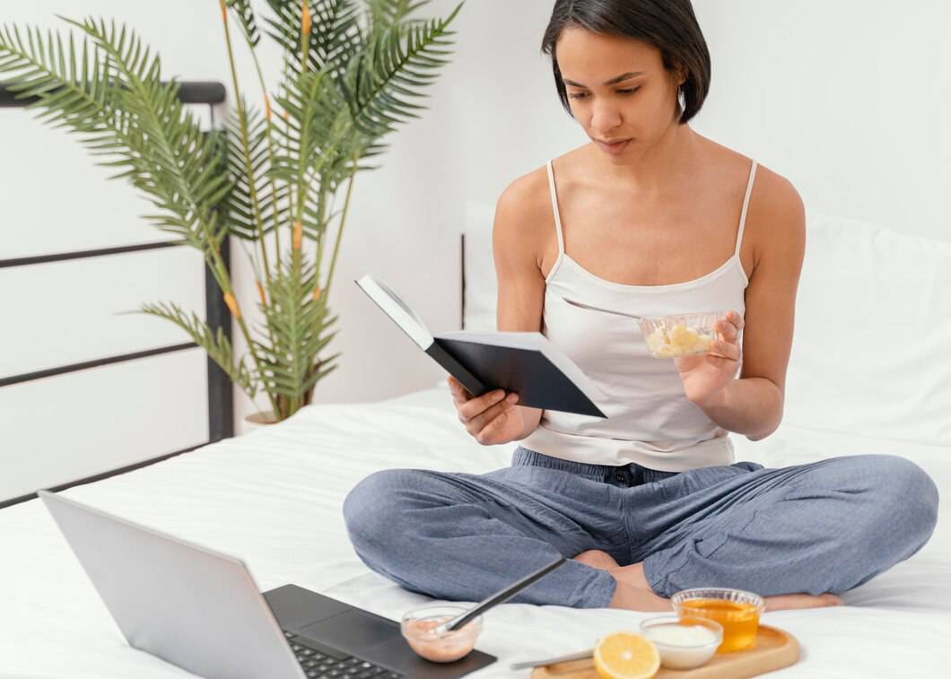 A person sits cross-legged on a bed, reading a book and enjoying a snack, with a laptop and snacks nearby, surrounded by greenery.