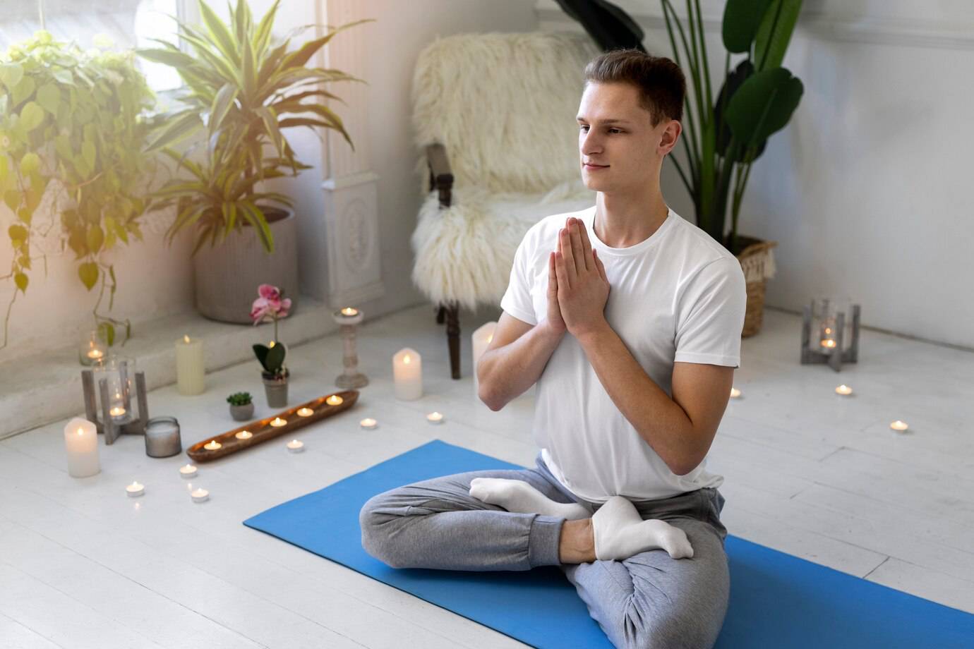 A person in a white shirt sits cross-legged on a blue yoga mat surrounded by plants and candles in a calming indoor space.