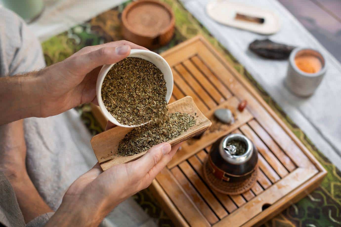 A person's hands pour loose herbs from a bowl onto a wooden dish, with a tea set and decorative mat visible in the background.