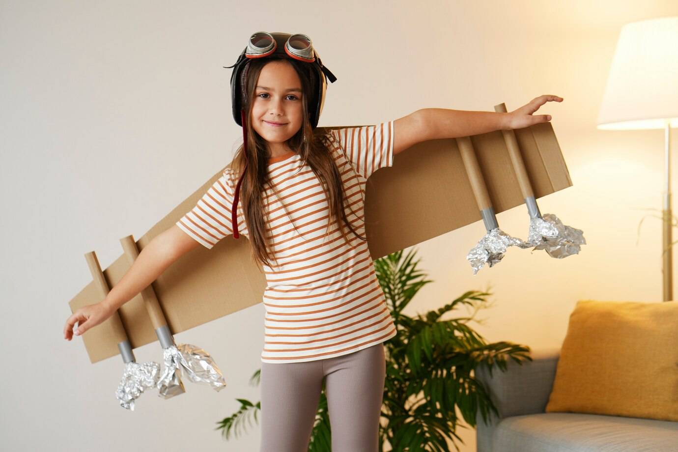A child wearing a striped shirt and a pilot's helmet poses with cardboard wings, showcasing imagination and creativity in a cozy room.