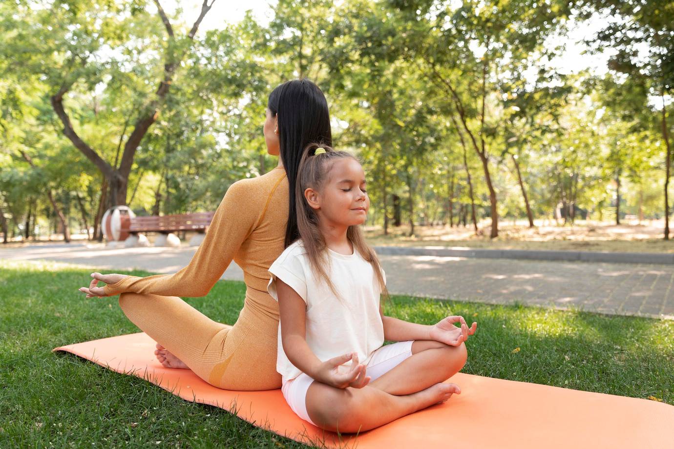 A woman and a child practice yoga on an orange mat in a lush green park, surrounded by trees and sunlight.