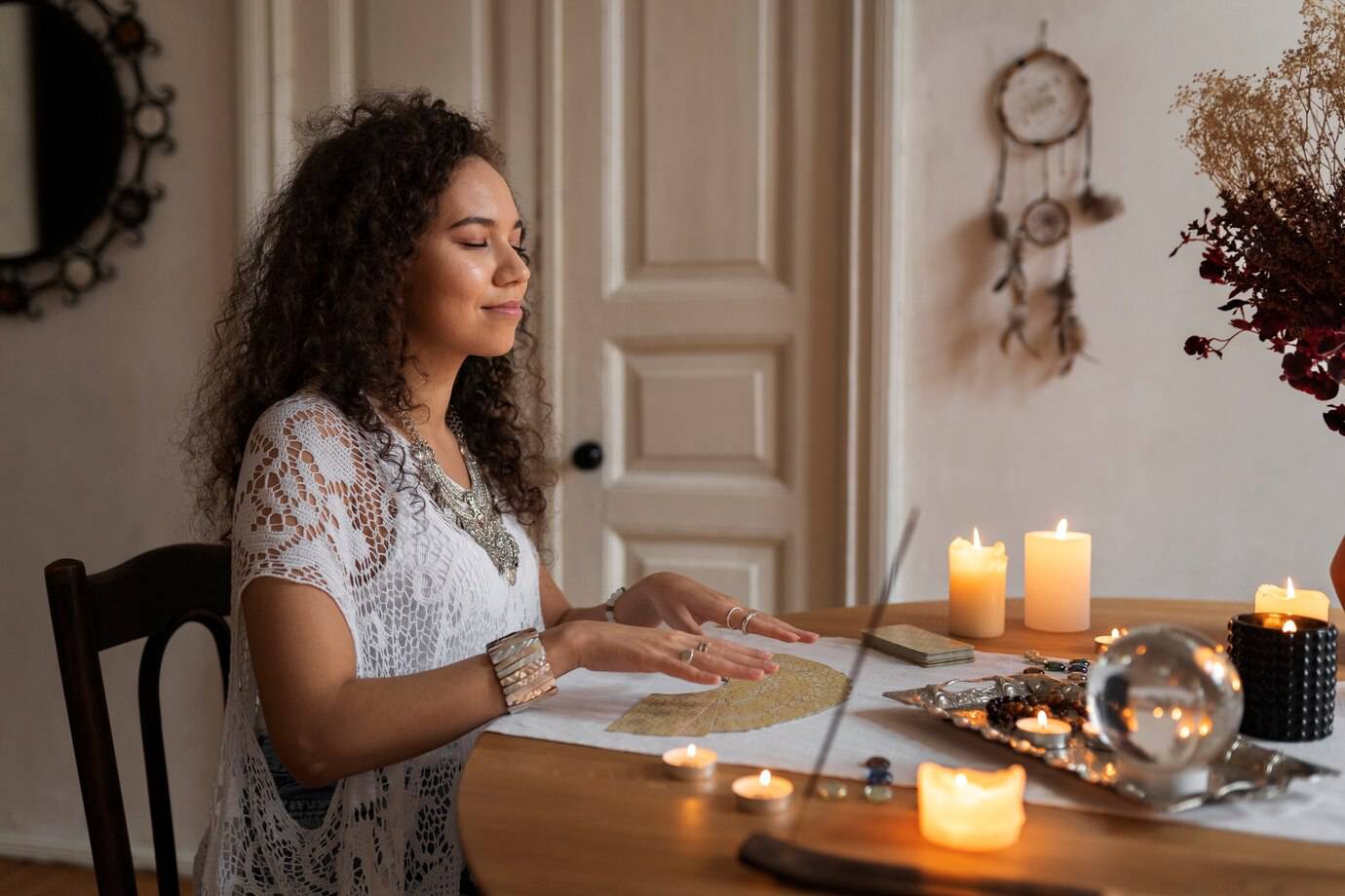 A woman in a bohemian-style outfit sits at a table adorned with candles, crystals, and a crystal ball, creating a mystical atmosphere.