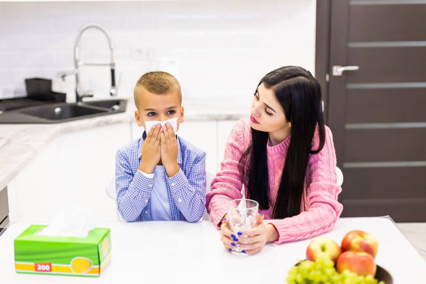 A child in a blue checkered shirt holds their hands to their face, while a woman in a pink sweater holds a glass of water at a kitchen table.