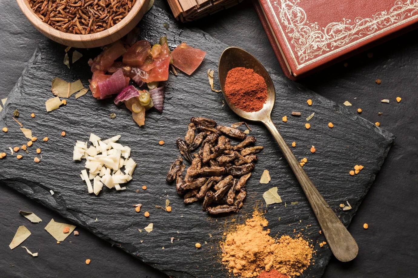 A slate board featuring various ingredients: edible insects, diced vegetables, spices, and a decorative book in the background.