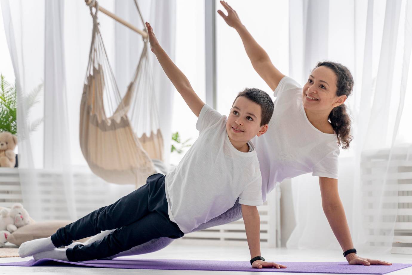 A woman and child practicing yoga together in a bright room, demonstrating the side plank pose on a purple mat.