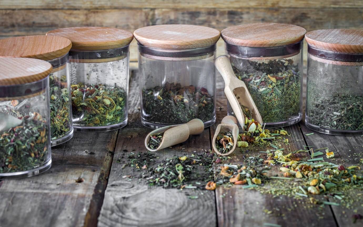 A collection of clear jars filled with various dried herbs, accompanied by a wooden scoop and scattered herbs on a rustic wooden surface.