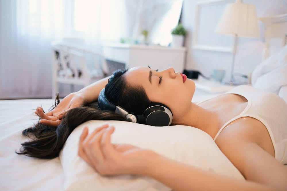 A woman lies on a bed wearing headphones, relaxing with arms outstretched, in a bright, softly lit room.