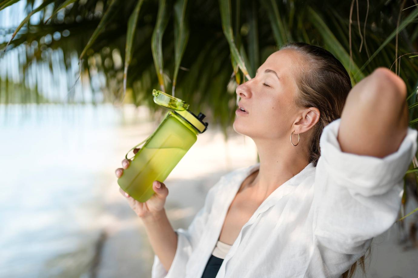 A person in a white shirt holds a green water bottle, enjoying a sunny day by the beach beneath tropical foliage.