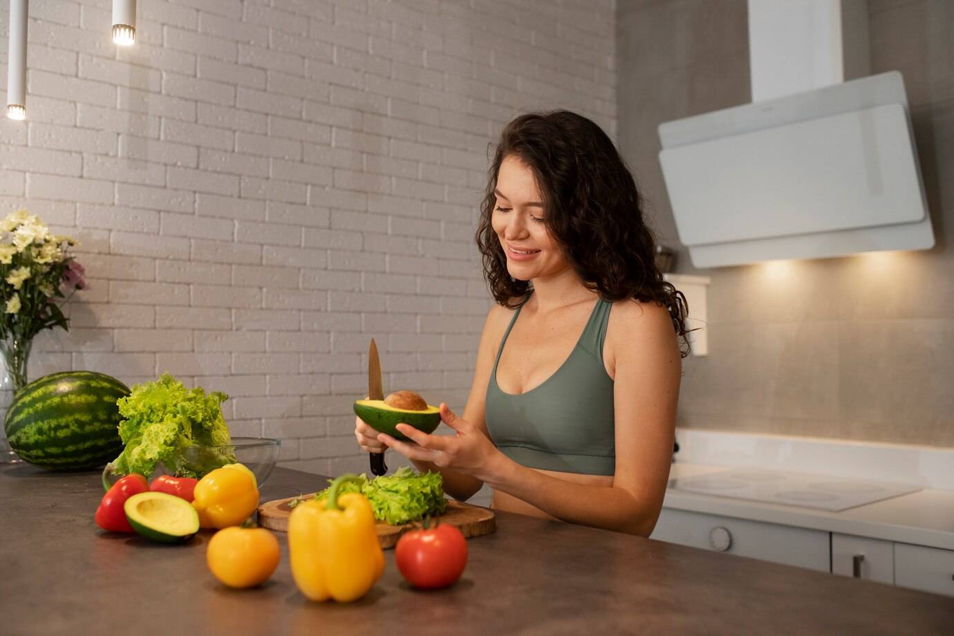 A woman in a kitchen preparing a salad, surrounded by fresh vegetables like lettuce, tomatoes, peppers, and a watermelon.