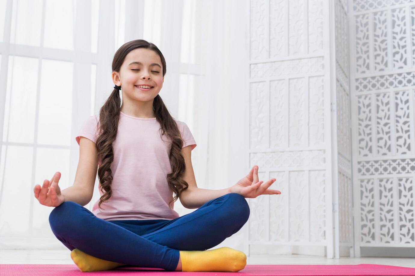 A child practices yoga in a serene indoor space, sitting cross-legged on a pink mat, with hands in a meditative position.