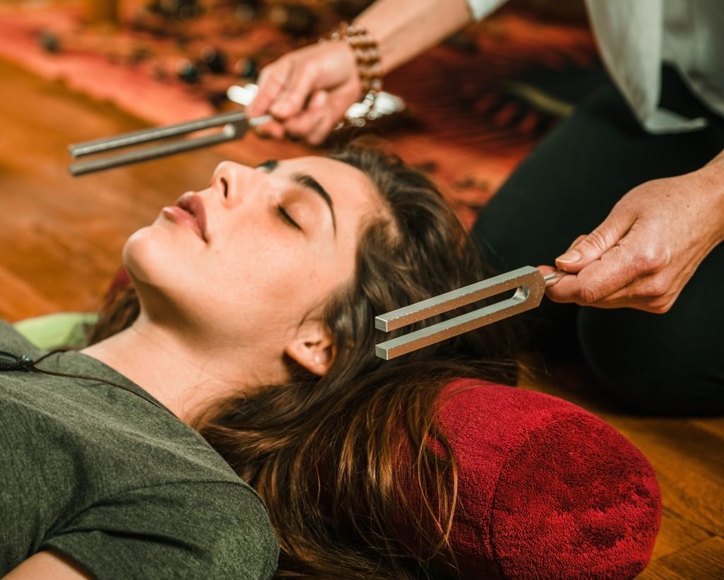 A person lies on a mat with a red bolster, while another individual uses tuning forks for sound therapy. Natural light fills the cozy space.