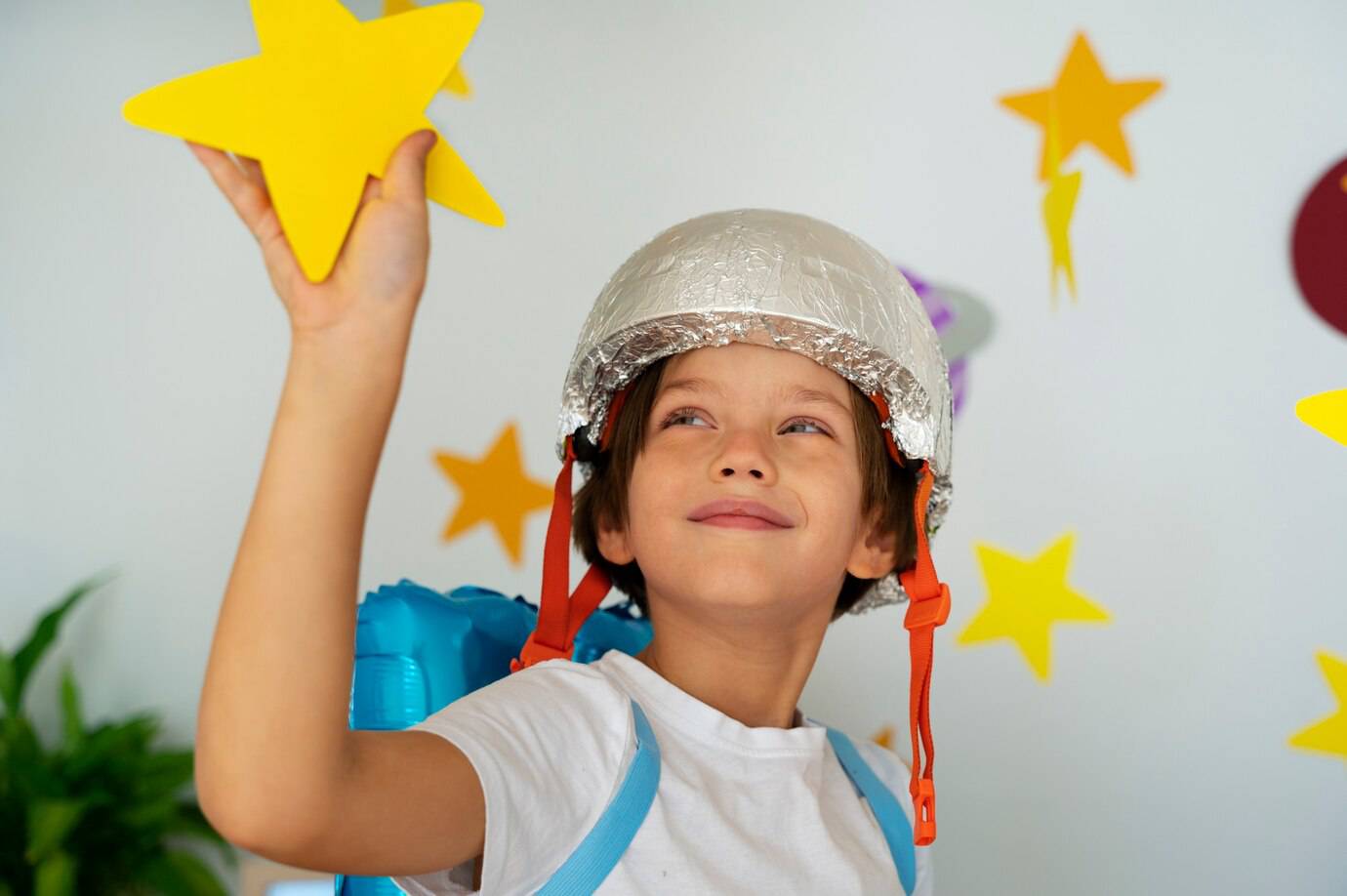 A child wearing a foil helmet and a blue backpack, joyfully holding a yellow star against a decorated wall.
