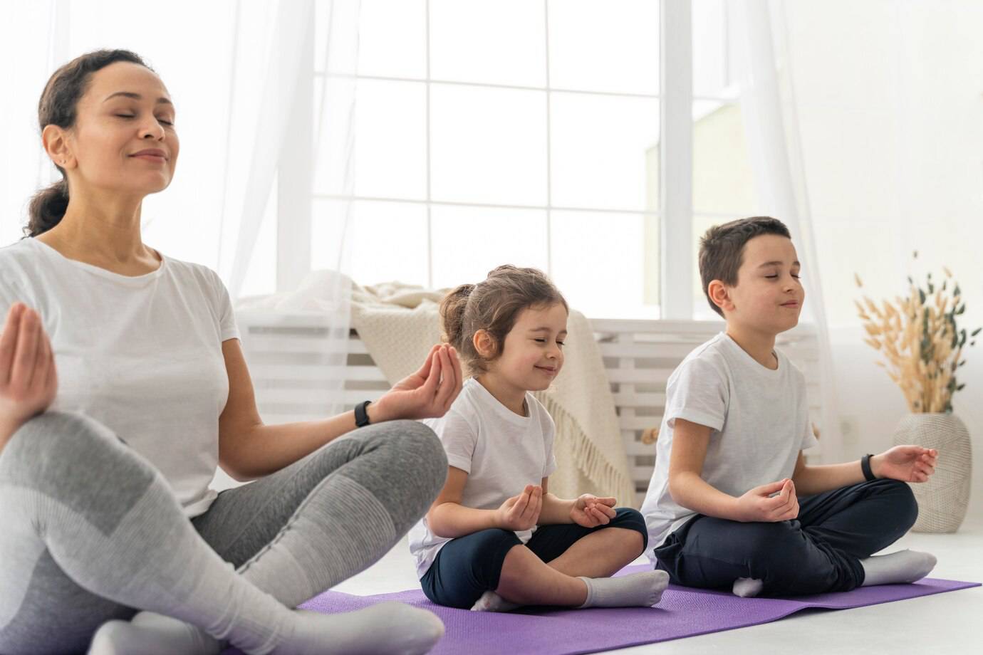 A woman and two children sit in a serene room, practicing meditation on purple yoga mats, surrounded by natural light and soft decor.