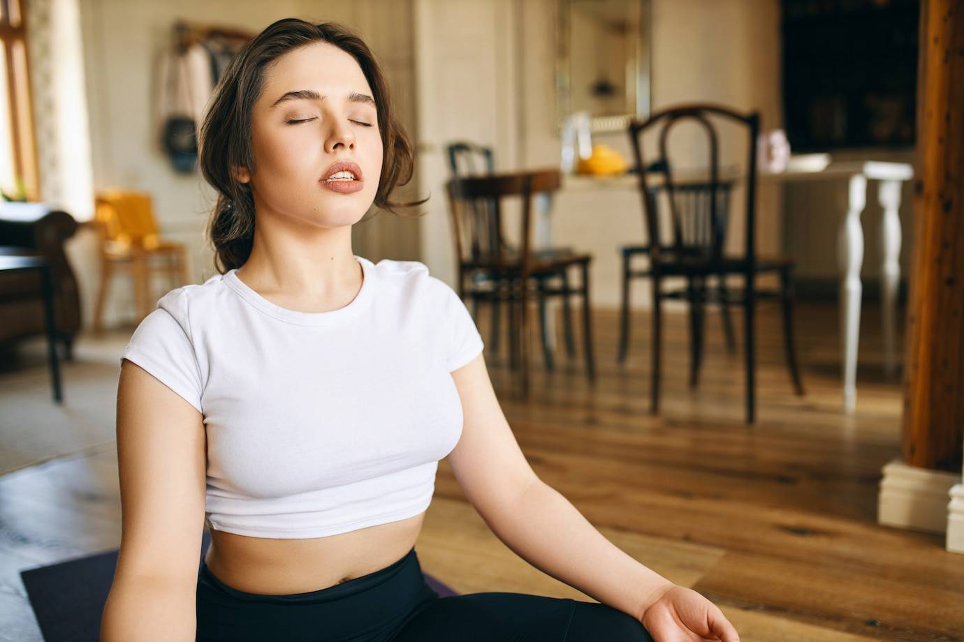 A woman sits cross-legged on a yoga mat in a cozy room, practicing mindfulness amidst wooden furniture and natural light.