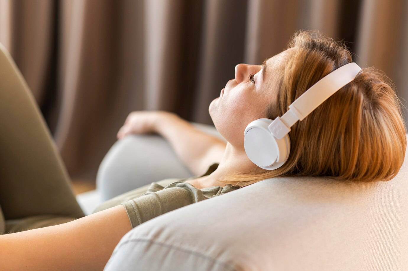A woman with headphones relaxes on a couch, eyes closed, enjoying music in a cozy, softly lit room.