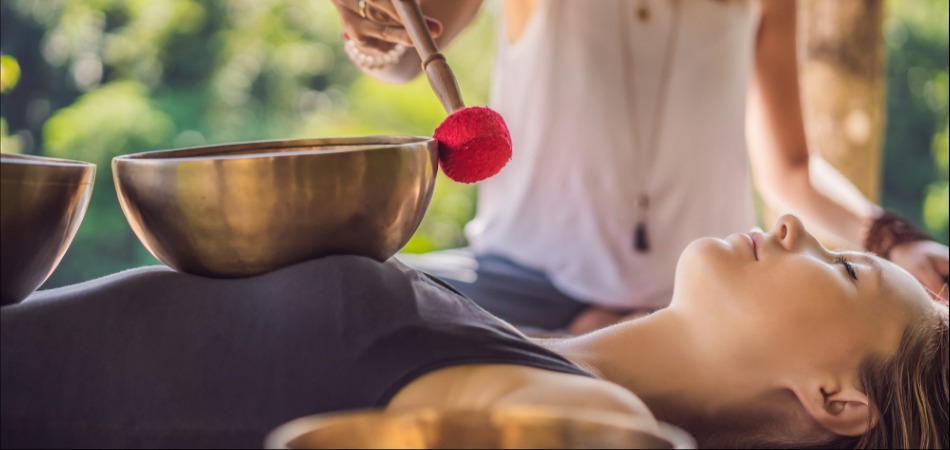 A practitioner uses a red mallet to strike a singing bowl, creating soothing sounds for a person lying on a massage table in a serene outdoor setting.