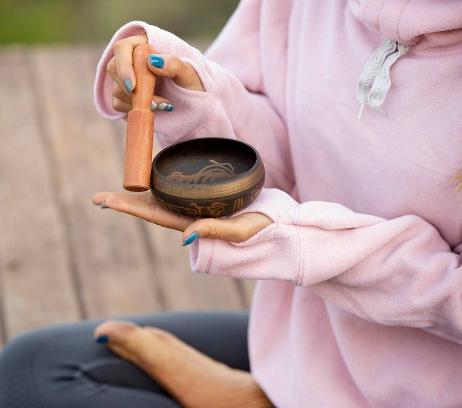 A person in a pink hoodie holds a wooden mallet above a decorative singing bowl, sitting in a serene outdoor setting.