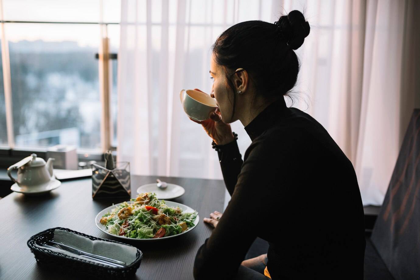 A person sitting at a table with a bowl, enjoying a fresh salad with colorful vegetables, and a teapot nearby, overlooking a city view.