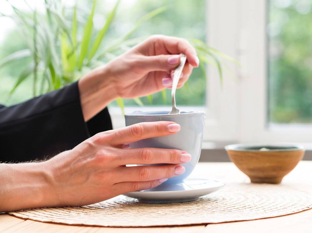 A person stirs a light blue cup with a spoon, sitting at a wooden table surrounded by green plants and natural light.