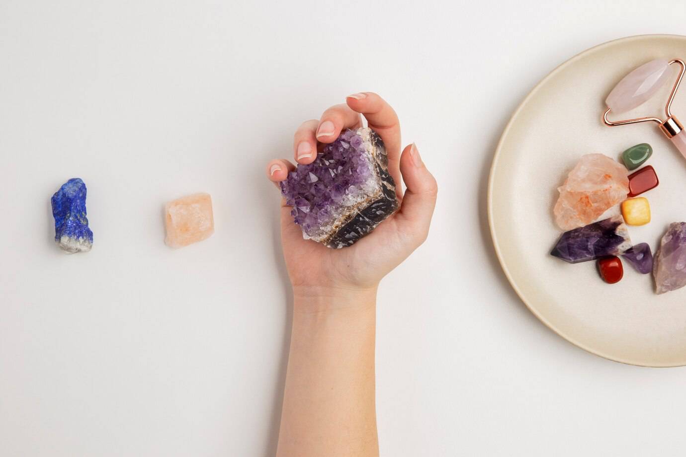 A hand holds a large amethyst crystal, surrounded by smaller crystals and a plate of various colorful stones and a rose quartz roller.