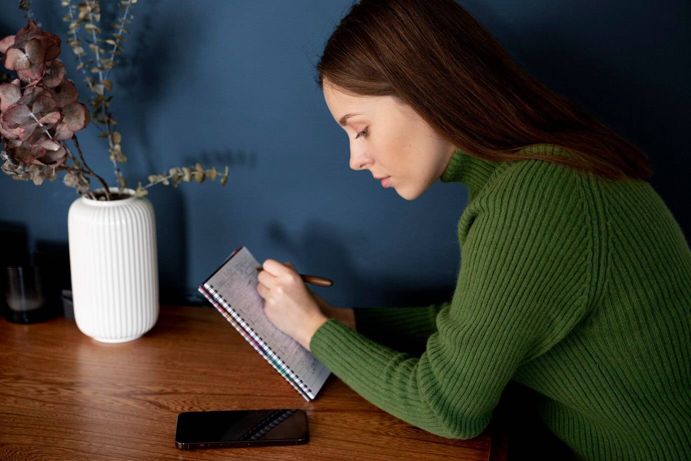 A person writing in a notebook with a pen, sitting at a wooden table adorned with a white vase and a smartphone nearby.