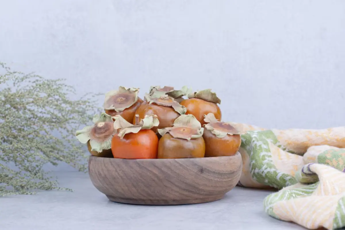 A wooden bowl filled with ripe persimmons and decorative leaves, placed next to a soft, patterned fabric and a delicate plant.